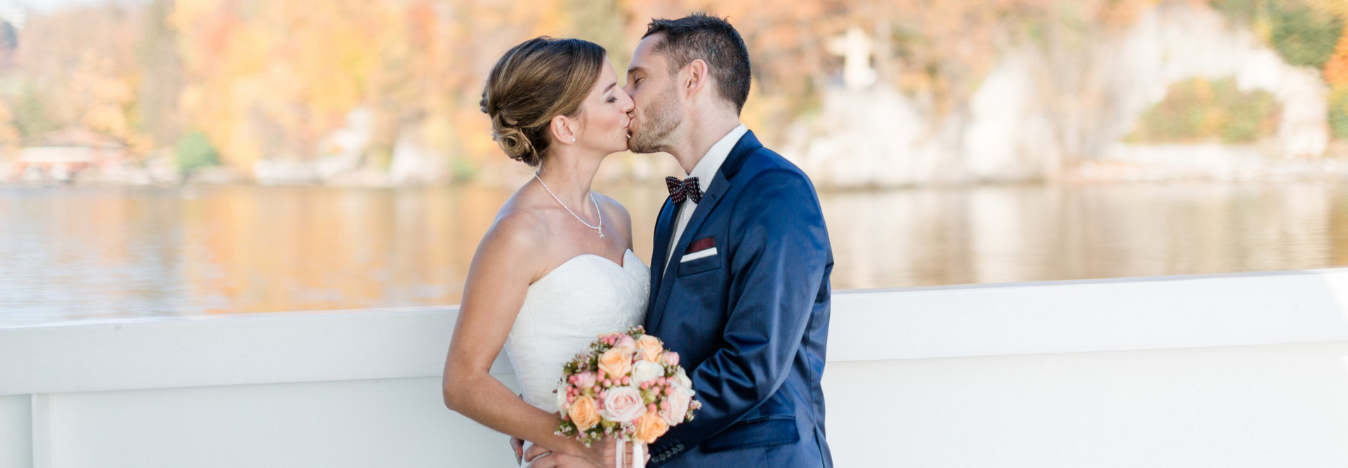 Couple de mariés s'embrassant dehors sur le bateau. En arrière-plan, on peut voir le lac et la forêt aux feuilles colorées, floues.