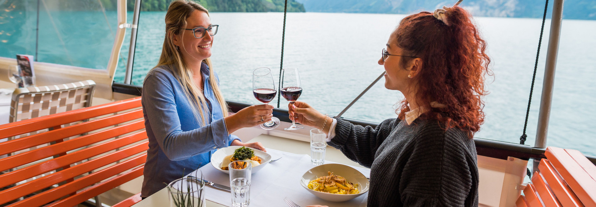 Zwei junge Frauen geniessen das Gastronomie Angebot auf dem Schiff und stossen mit einem Glas Rotwein an.